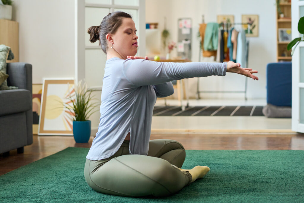 Side view of girl with Down syndrome sitting on the floor with crossed legs while practicing yoga exercise or position in living room