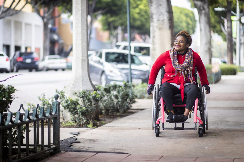 A young black disabled woman with a wheelchair and a bright colored sweater on a city sidewalk.