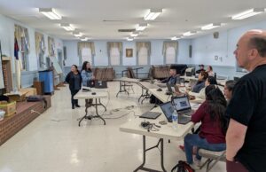 People seated at tables in a room while on computers. Two instructors stand at the front of the room. A man stands to the right of the frame.
